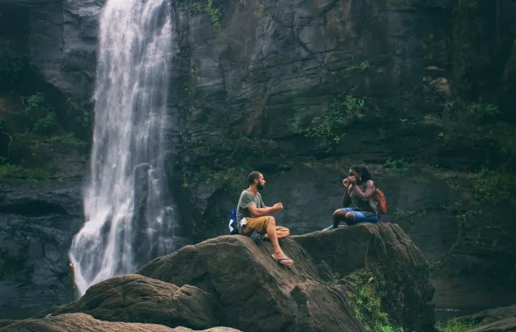 Two people seated on a rock by a waterfall