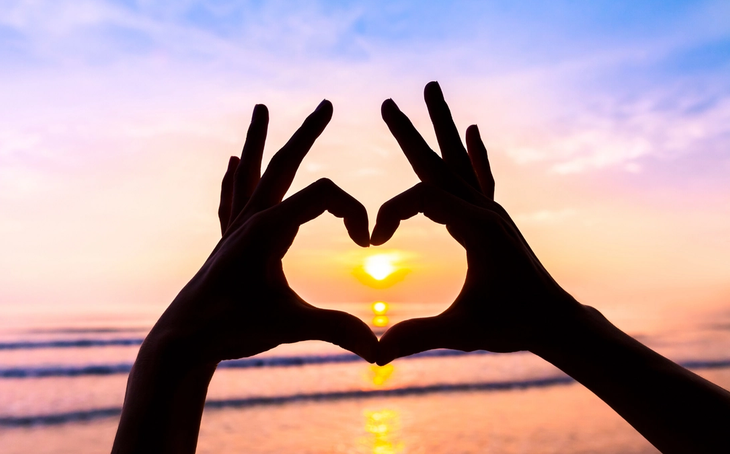 A person make a heart shape with her fingers in front of a sunset
