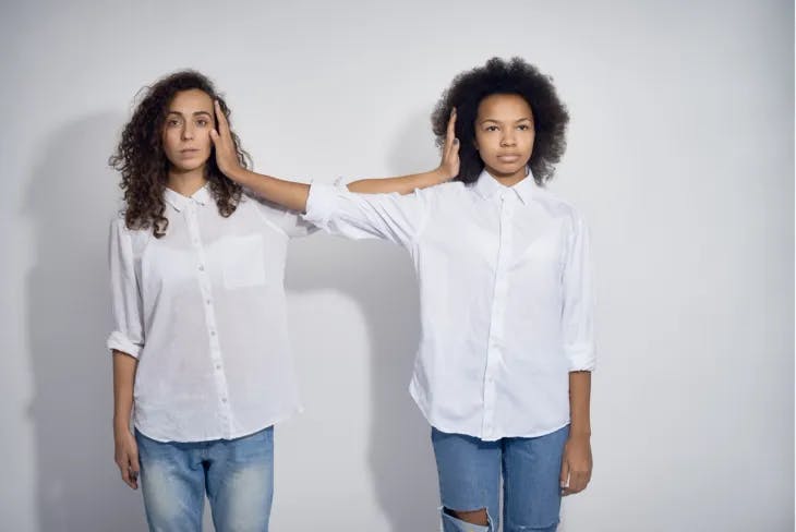 Two girls standing side by side, extending their arms to touch each other's faces